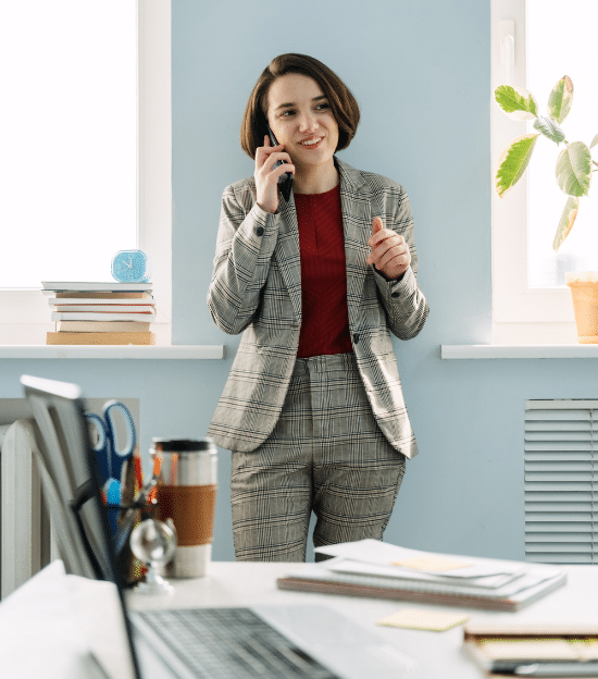 An attorney is seen smiling on a phone standing in front of a desk in a bright office with a blue wall representing Temporary Legal Staffing