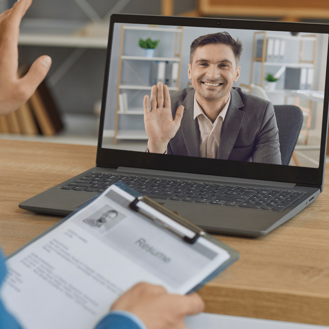 The 7-Point Checklist for Conducting a Successful Virtual Interview - male recruiter sitting at desk with resume in his hand waving at male job candidate on a laptop screen