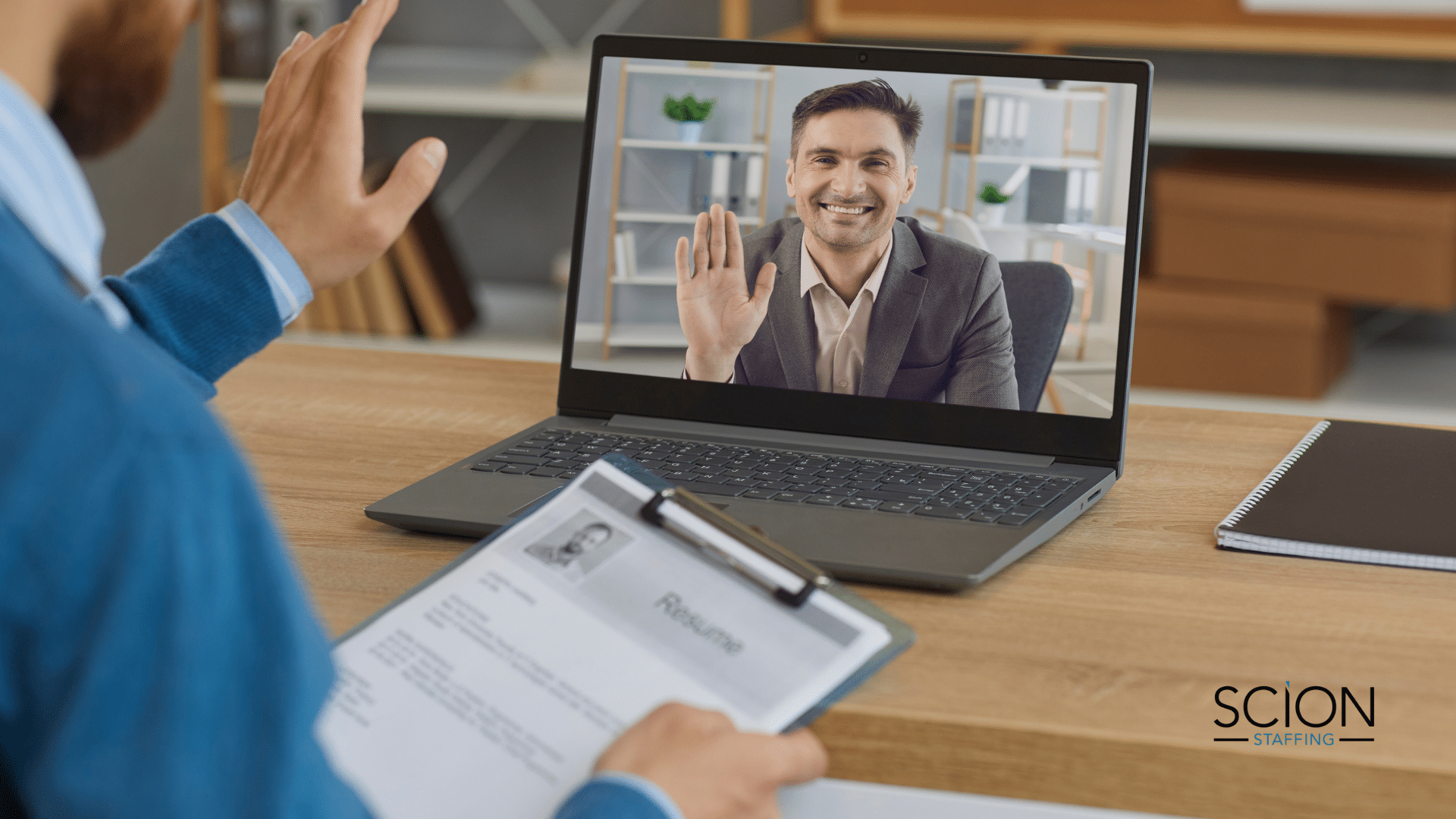 The 7-Point Checklist for Conducting a Successful Virtual Interview - male recruiter sitting at desk with resume in his hand waving at male job candidate on a laptop screen